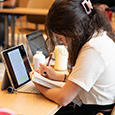 A student works with a laptop, a stack of papers, and a pen in the library