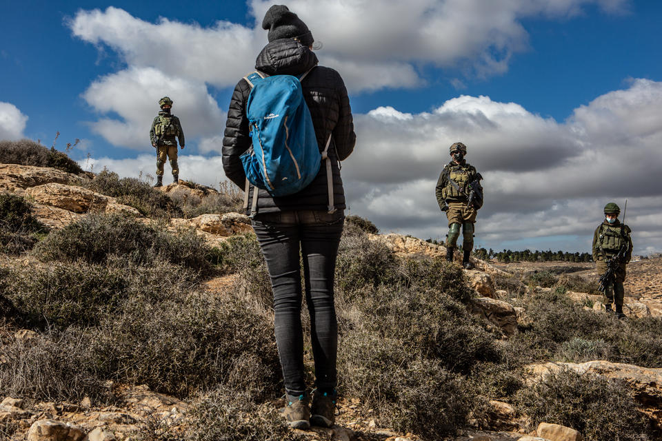 A figure in a black coast and with a blue backpack stands facing a hillside with three Israeli soldiers