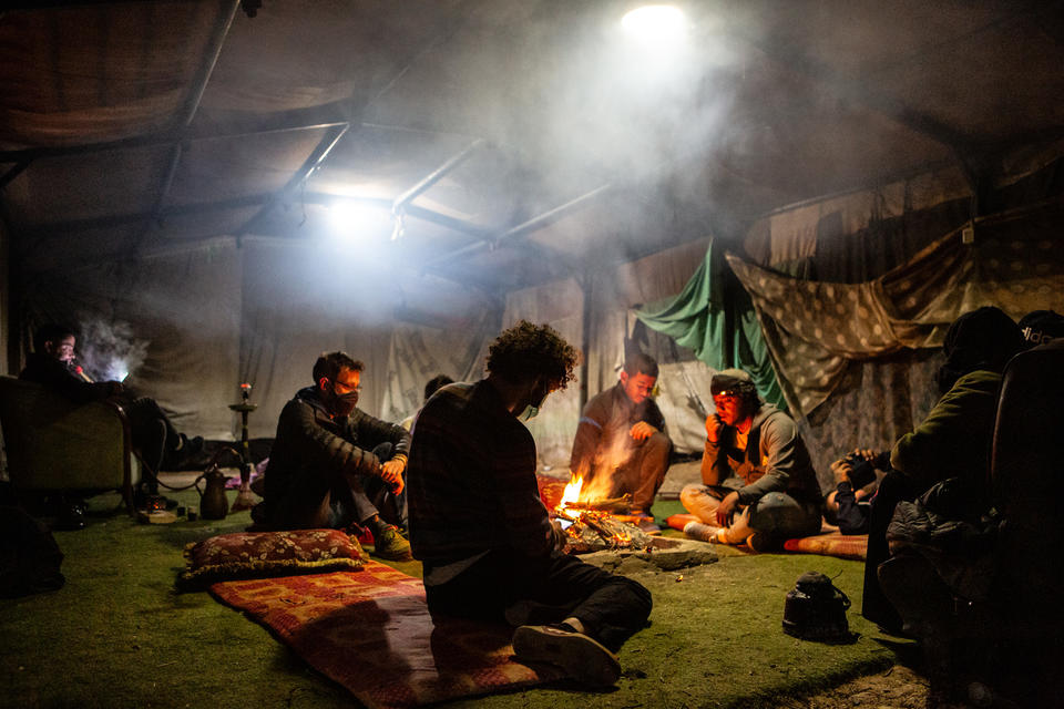 five figure sit around a small fire, smoke rises to two lights hanging overhead