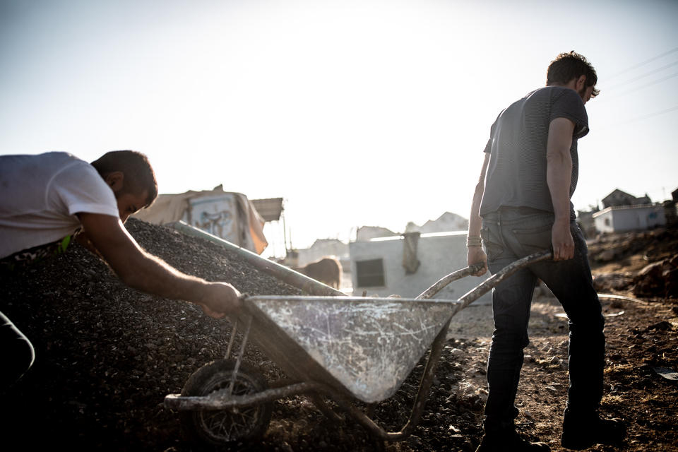 two men in t-shirts carry a metal wheelbarrow near a pile of gravel