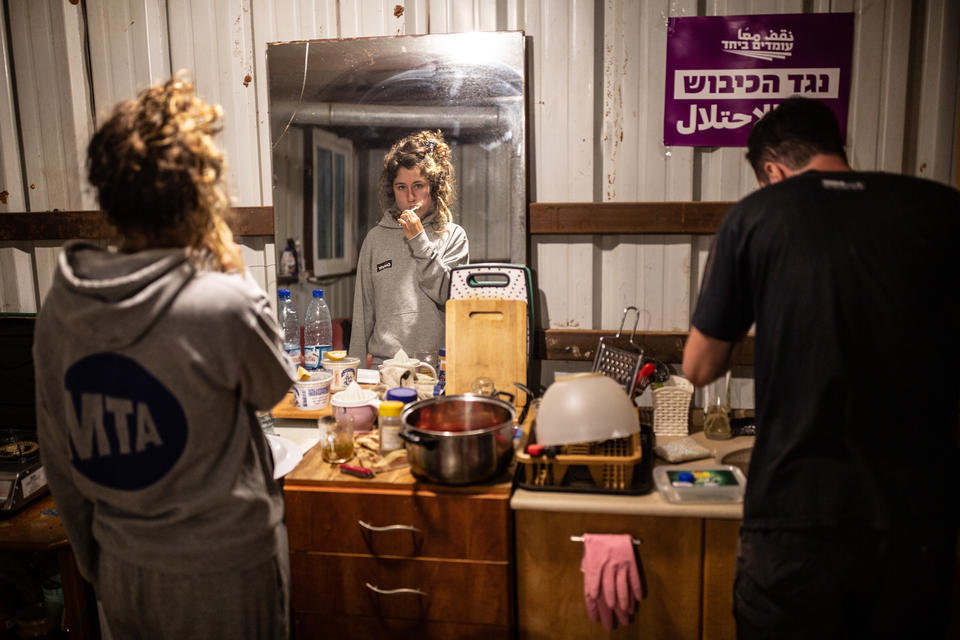 A woman brushes her teeth gazing into a mirror next to someone doing dishes